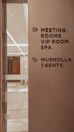A corridor in a building with signage indicating directions to various facilities including meeting rooms, a VIP room, a spa, a Musholla, and a men's restroom. The wall is light-colored with dark brown text and icons. The area is well-lit with reflective surfaces and marble flooring.
