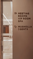 A corridor in a building with signage indicating directions to various facilities including meeting rooms, a VIP room, a spa, a Musholla, and a men's restroom. The wall is light-colored with dark brown text and icons. The area is well-lit with reflective surfaces and marble flooring.