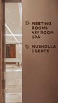 A corridor in a building with signage indicating directions to various facilities including meeting rooms, a VIP room, a spa, a Musholla, and a men's restroom. The wall is light-colored with dark brown text and icons. The area is well-lit with reflective surfaces and marble flooring.