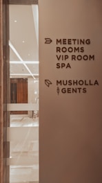 A corridor in a building with signage indicating directions to various facilities including meeting rooms, a VIP room, a spa, a Musholla, and a men's restroom. The wall is light-colored with dark brown text and icons. The area is well-lit with reflective surfaces and marble flooring.