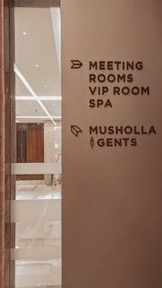 A corridor in a building with signage indicating directions to various facilities including meeting rooms, a VIP room, a spa, a Musholla, and a men's restroom. The wall is light-colored with dark brown text and icons. The area is well-lit with reflective surfaces and marble flooring.