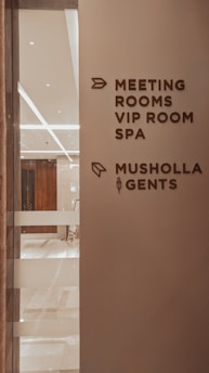 A corridor in a building with signage indicating directions to various facilities including meeting rooms, a VIP room, a spa, a Musholla, and a men's restroom. The wall is light-colored with dark brown text and icons. The area is well-lit with reflective surfaces and marble flooring.