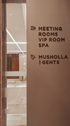 A corridor in a building with signage indicating directions to various facilities including meeting rooms, a VIP room, a spa, a Musholla, and a men's restroom. The wall is light-colored with dark brown text and icons. The area is well-lit with reflective surfaces and marble flooring.