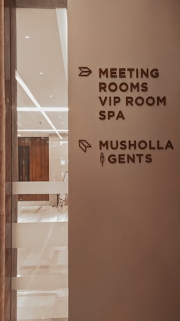 A corridor in a building with signage indicating directions to various facilities including meeting rooms, a VIP room, a spa, a Musholla, and a men's restroom. The wall is light-colored with dark brown text and icons. The area is well-lit with reflective surfaces and marble flooring.