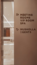 A corridor in a building with signage indicating directions to various facilities including meeting rooms, a VIP room, a spa, a Musholla, and a men's restroom. The wall is light-colored with dark brown text and icons. The area is well-lit with reflective surfaces and marble flooring.