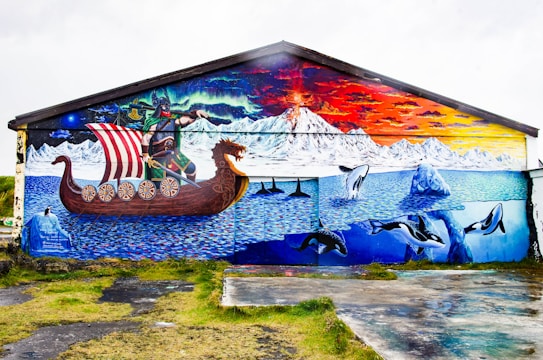A vibrant mural depicts a scene with a Viking ship sailing in icy waters. A warrior stands confidently on the ship as orcas leap through the waves. Snow-capped mountains and an aurora borealis light up the sky, creating a dramatic contrast with the sunset.