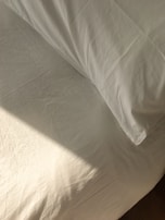 A neatly made bed with white sheets and a pillow is bathed in soft, natural light. The fabric has a slight texture and wrinkles, hinting at the presence of sunlight casting subtle shadows.