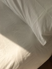 A neatly made bed with white sheets and a pillow is bathed in soft, natural light. The fabric has a slight texture and wrinkles, hinting at the presence of sunlight casting subtle shadows.