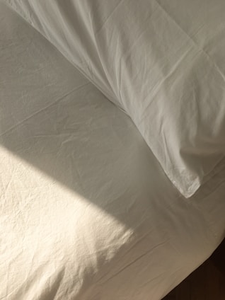 A neatly made bed with white sheets and a pillow is bathed in soft, natural light. The fabric has a slight texture and wrinkles, hinting at the presence of sunlight casting subtle shadows.