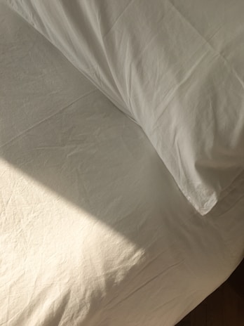 A neatly made bed with white sheets and a pillow is bathed in soft, natural light. The fabric has a slight texture and wrinkles, hinting at the presence of sunlight casting subtle shadows.
