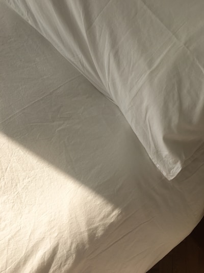 A neatly made bed with white sheets and a pillow is bathed in soft, natural light. The fabric has a slight texture and wrinkles, hinting at the presence of sunlight casting subtle shadows.