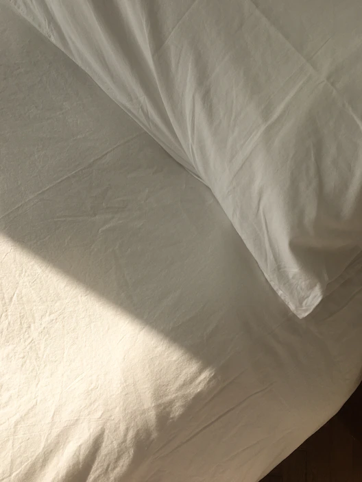 A neatly made bed featuring soft, pastel-colored sheets and fluffy pillows bathed in morning sunlight.