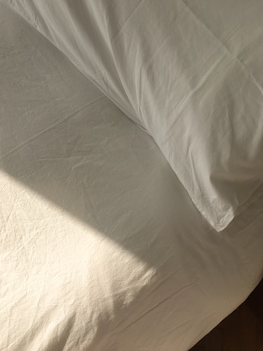 A neatly made bed with white sheets and a pillow is bathed in soft, natural light. The fabric has a slight texture and wrinkles, hinting at the presence of sunlight casting subtle shadows.