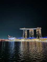A night view of a waterfront scene featuring a distinctive, architecturally unique hotel with three tall towers connected by a rooftop structure resembling a ship. Adjacent to it is a futuristic-looking building with a design reminiscent of a lotus flower. The structures are illuminated with bright lights, reflecting on the calm water below.
