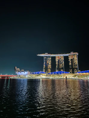A night view of a waterfront scene featuring a distinctive, architecturally unique hotel with three tall towers connected by a rooftop structure resembling a ship. Adjacent to it is a futuristic-looking building with a design reminiscent of a lotus flower. The structures are illuminated with bright lights, reflecting on the calm water below.