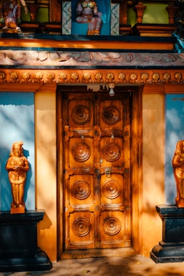 A beautifully ornate wooden door, featuring intricate circular carvings, is flanked by two golden statues mounted on black pedestals. There are additional colorful details like blue and yellow painted walls, and figurines positioned above the door as part of the structure.