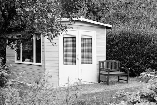 A small wooden shed with a window and a door, surrounded by a garden. There is a wooden bench situated in front of the shed, and dense foliage can be seen in the background.