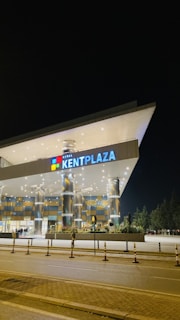 A modern, illuminated shopping mall entrance with reflective columns and a prominent sign displaying 'Konya Kentplaza'. The building is well-lit against the night sky, and there are bollards and a road in the foreground.