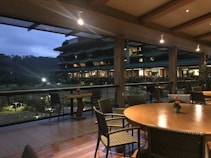 An outdoor seating area features wooden tables and woven chairs under a covered terrace. Beyond the railing, a large building with multiple floors and illuminated windows is visible at dusk. The area has subtle lighting and small red flowers on the tables add a decorative touch.