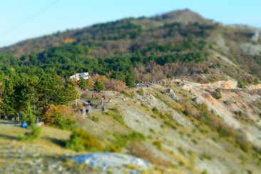 A scenic landscape features a hillside with numerous people walking along a path. The area is surrounded by lush green forests and rocky terrain. The sky is clear, suggesting a pleasant day.