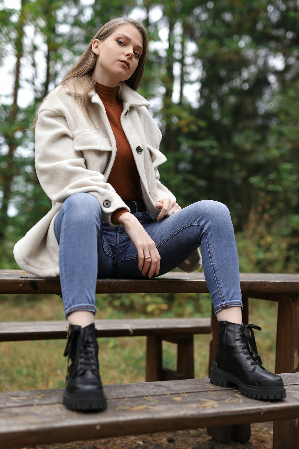 woman in beige coat and blue denim jeans sitting on brown wooden bench during daytime