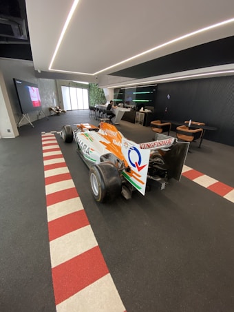 A Formula 1 car is displayed indoors on a floor with red and white striping. The car is primarily orange and white with sponsorship logos, including one that reads Kingfisher. The environment is modern with a seating area, a bar, and a large screen showing a video.