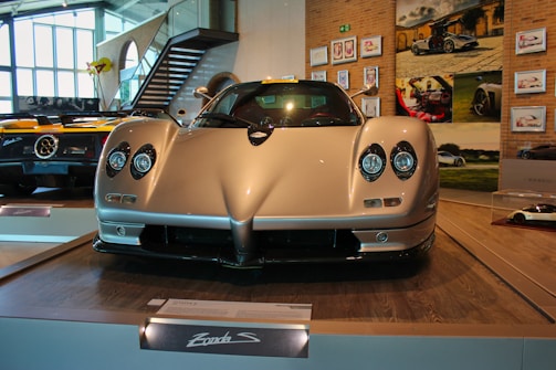 A sleek, high-performance sports car is displayed in a showroom with other luxury vehicles. The car features aerodynamic curves and distinctive headlights. The setting includes a stylish interior with art and photographs on the walls.