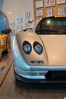 A close-up view of a sleek, silver sports car with prominent round headlights and smooth, aerodynamic design. The car is parked indoors, surrounded by framed pictures on the wall. Another sports car in yellow is visible to the side, and there is a plant in the background.
