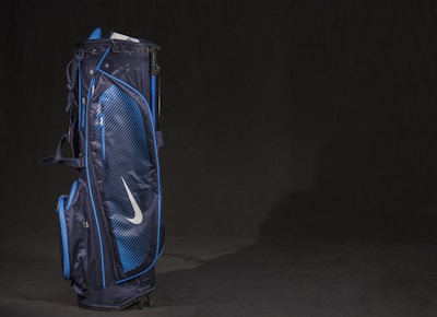 A dark blue golf bag with light blue accents and a prominent white swoosh logo. The design is sleek with multiple pockets and zippers visible, set against a plain dark background.