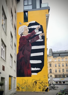 A large mural on the side of a building depicts a figure in historical clothing with a white wig and red coat, gesturing upward. The background is bright yellow with black and white stripes. Graffiti covers the lower part of the wall.
