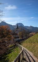 A peaceful rural scene featuring a church with a prominent steeple set against a backdrop of mountains. The pathway in the foreground curves gently, bordered by a wooden fence, and leads the eye towards the church. Trees with autumn foliage add a touch of color, and there are scattered houses near the church.