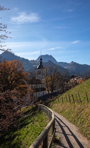 A peaceful rural scene featuring a church with a prominent steeple set against a backdrop of mountains. The pathway in the foreground curves gently, bordered by a wooden fence, and leads the eye towards the church. Trees with autumn foliage add a touch of color, and there are scattered houses near the church.