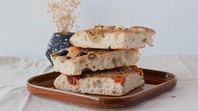 Artisanal focaccia with fresh herbs and olive oil, styled on a beige cotton paper background.