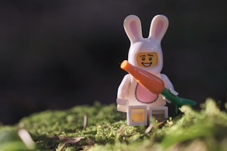 A small figurine dressed in a white bunny costume is holding an oversized carrot. It is standing on a patch of green moss, with soft sunlight highlighting its features. The background is blurred, creating a gentle contrast with the figure.