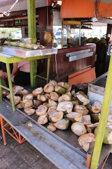 Coconuts are halved and stacked on a metal table at a street-side vendor. The background shows parts of a wooden stall and various equipment or storage areas. There are pieces of sugarcane on a tray above the coconuts. People are partially visible, sitting behind the table.