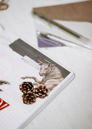 A notepad features an illustration of a bull alongside three pinecones. Pens and a folder are scattered on the light-colored surface around it.