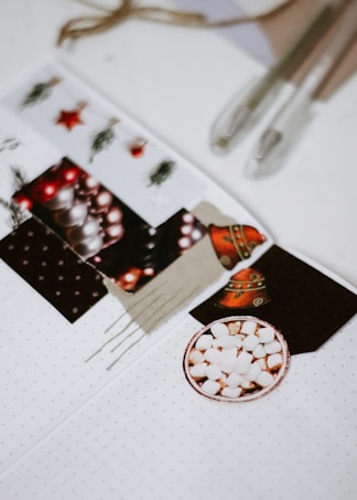 A collection of Christmas-themed cutouts is arranged on a white background, including images of ornaments, a decorated bell, and greenery. Two pens and a roll of twine are placed nearby, suggesting a crafting activity. A small round plate filled with marshmallows is prominently featured.