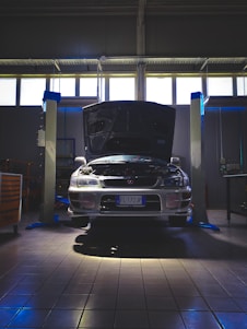 A car is positioned in a garage or workshop with its hood open, indicating it might be undergoing maintenance or inspection. The lighting is dim, with a few bright areas highlighting the vehicle. The space is industrial, with lift platforms and tool cabinets visible.