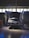 A car is positioned in a garage or workshop with its hood open, indicating it might be undergoing maintenance or inspection. The lighting is dim, with a few bright areas highlighting the vehicle. The space is industrial, with lift platforms and tool cabinets visible.