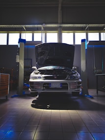 A car is positioned in a garage or workshop with its hood open, indicating it might be undergoing maintenance or inspection. The lighting is dim, with a few bright areas highlighting the vehicle. The space is industrial, with lift platforms and tool cabinets visible.