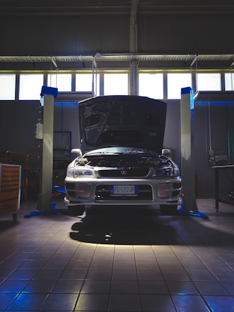 A car is positioned in a garage or workshop with its hood open, indicating it might be undergoing maintenance or inspection. The lighting is dim, with a few bright areas highlighting the vehicle. The space is industrial, with lift platforms and tool cabinets visible.
