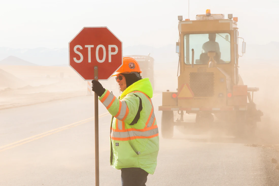 Traffic controller in hi-vis holding a stop bat in front of a grader