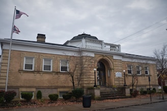 A historic brick building with a domed roof and adorned with white pillars and trim. The entrance features a prominent archway with steps leading up to it. Two flags are flying on flagpoles, and there are trees and shrubs in a landscaped area surrounding the sidewalk in front.