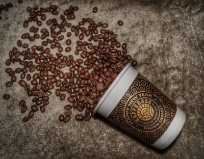A takeaway coffee cup is tilted on its side, allowing numerous coffee beans to spill out onto a textured surface. The cup features a decorative pattern with words like 'KAFFEBASE,' 'HAPPY NEW YEAR,' and 'HAPPY DIWALI.' The coffee beans are scattered around the cup, creating an artistic and inviting scene.