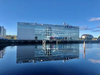 A sleek glass office building reflecting the nearby river under a clear sky.