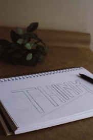 A spiral-bound sketchbook lies open on a brown surface, displaying a pencil outline of the word 'THINK' in large block letters. A pencil rests beside the notebook. To the left, there is a small plant with dark green leaves partially blurred in the background.