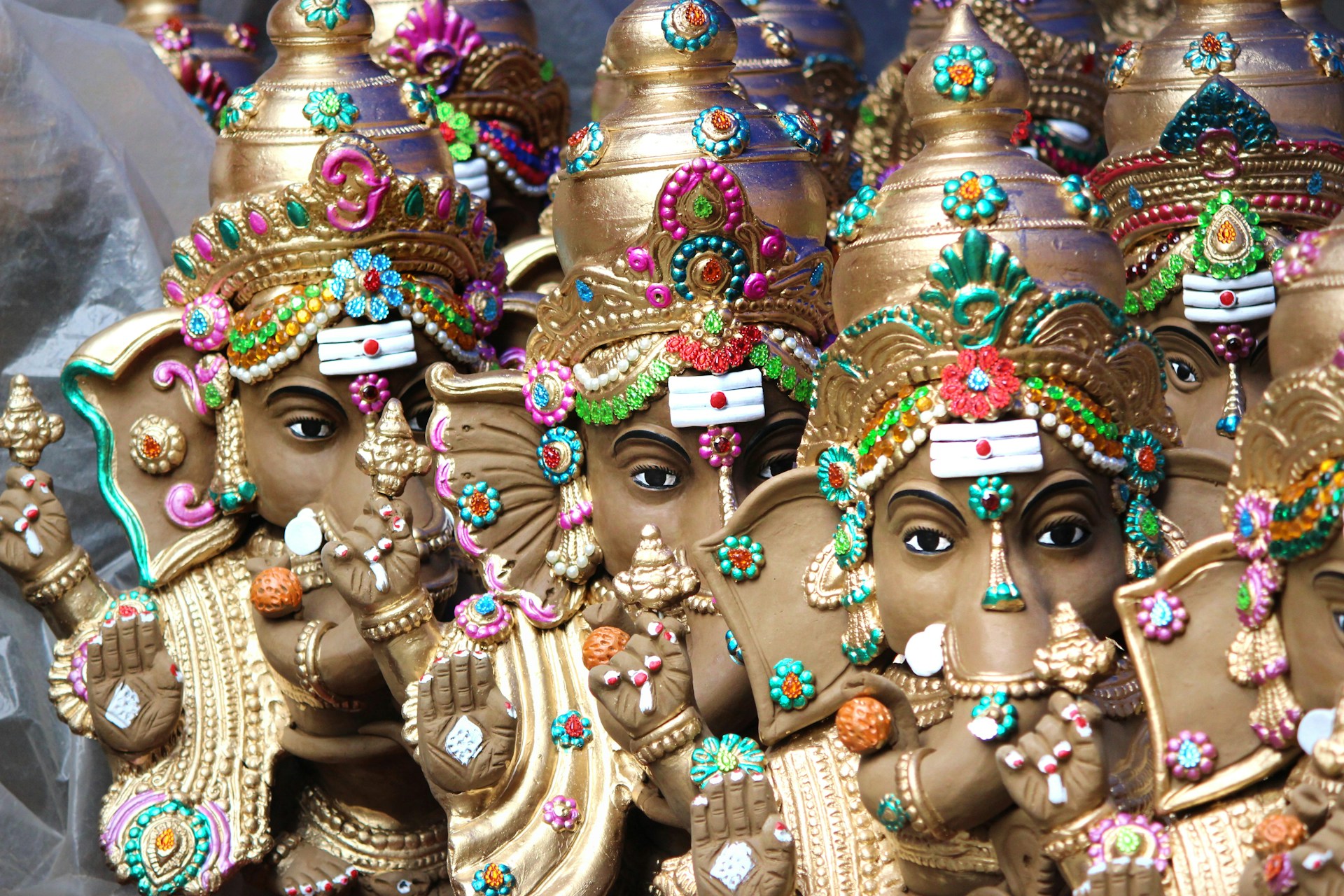A collection of intricately crafted statues depicting an elephant-headed deity adorned with colorful ornate jewelry and headdresses. Each statue features vibrant embellishments with beads and jewels in various colors, displaying ceremonial poses and expressions.