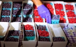 A gloved hand is carefully selecting a container of blueberries from a display of fruit. The arrangement consists of multiple rectangular cartons filled with ripe blueberries and vibrant red raspberries in a well-organized pattern. The blueberries and raspberries are held in blue cardstock containers, typical at farmer's markets, against a floral and checkered tablecloth backdrop.