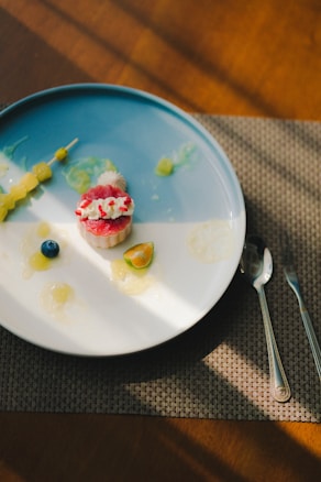 A minimalistic plate of food is arranged on a round blue and white plate. The plate contains a small dessert topped with white and red cream, a sliced piece of citrus fruit, a single blueberry, and a small skewer of yellow grapes. The plate sits on a woven placemat with two silver utensils beside it. There are soft shadows cast across the table by sunlight.