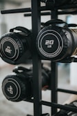 Several cylindrical fitness sandbags are stored on a black metal rack. The visible sandbags are labeled with different weights such as 20, 30, and 40 pounds and feature handles for easy carrying.